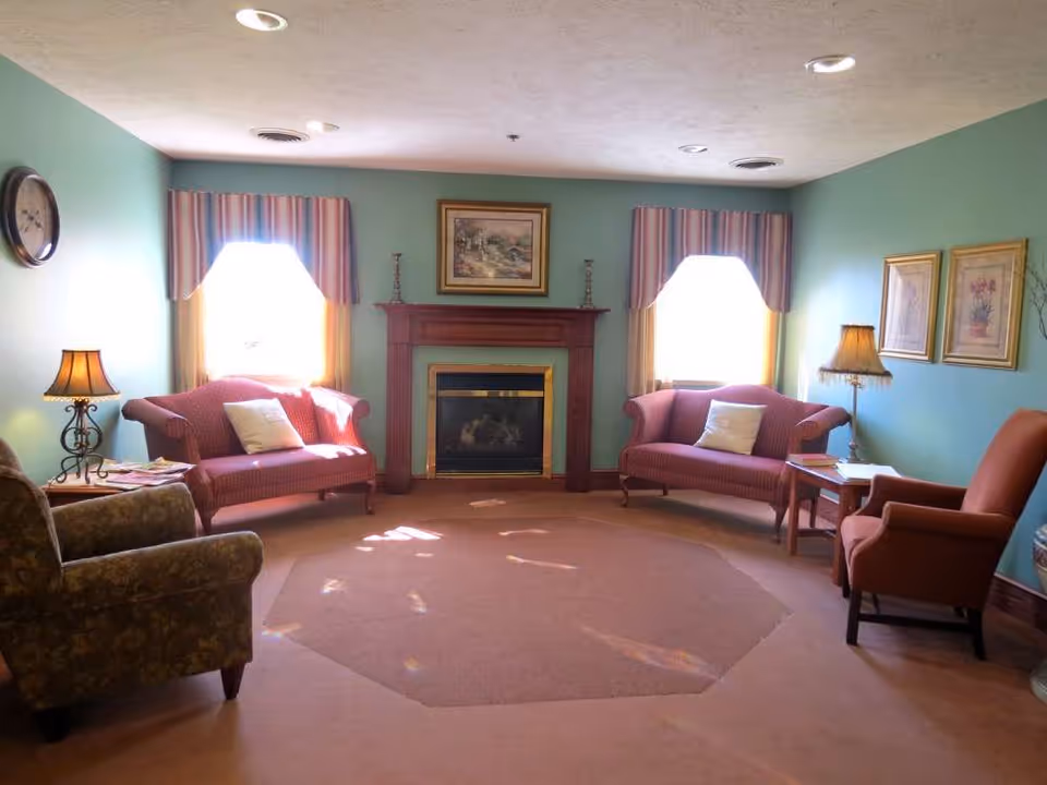 A cozy living room with two red upholstered loveseats, a green patterned armchair, and a red armchair arranged around a fireplace with a wooden mantel. The walls are painted green and decorated with framed artwork and a clock. Two windows with striped valances allow natural light into the room, and there are two table lamps providing additional lighting.