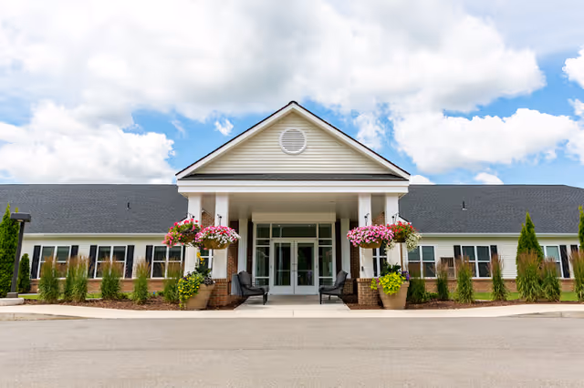 Front exterior view of StoryPoint Rockford facility with a covered entrance, two chairs, hanging flower baskets, and potted plants under a partly cloudy sky.