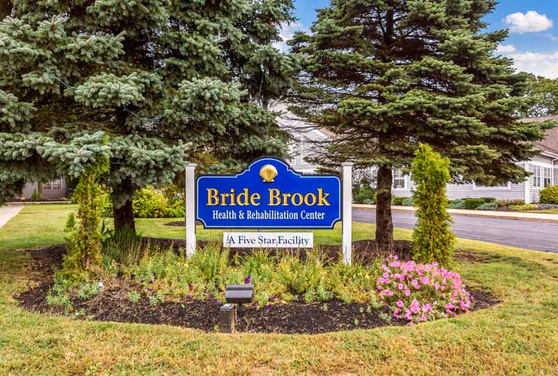 A blue sign reading "Bride Brook Health & Rehabilitation Center — A Five Star Facility" stands in a landscaped flowerbed with evergreen trees and a driveway behind it.