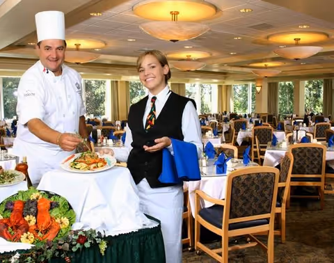 A chef and a waitress standing in a dining room of a senior living facility. The chef is holding a plate of food and garnishing it, while the waitress smiles and holds a blue napkin. The dining room is set with tables covered in white tablecloths, blue napkins, and chairs, with large windows letting in natural light.