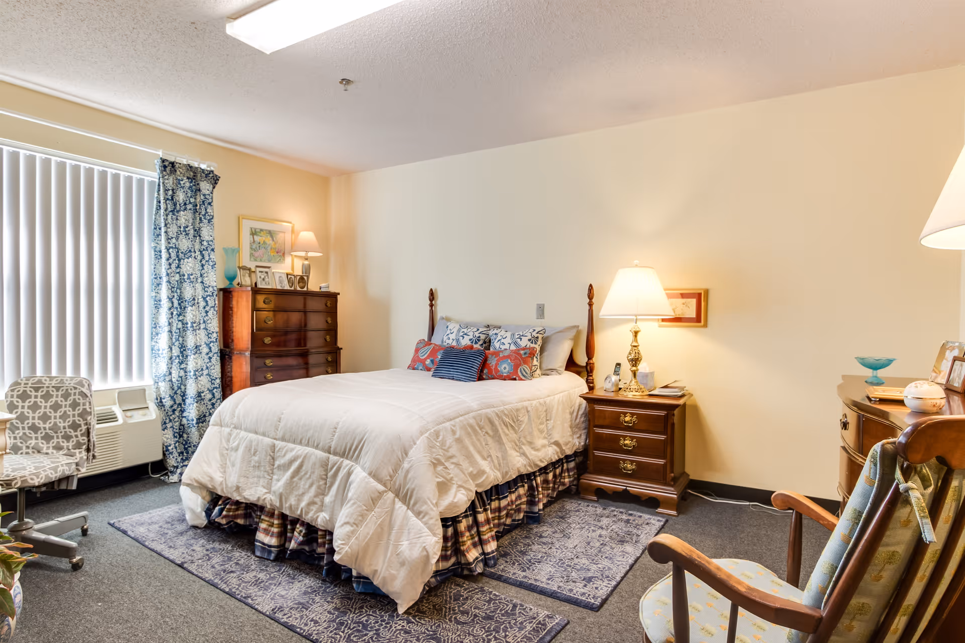 A cozy bedroom in an assisted living facility featuring a neatly made bed with white bedding and multiple decorative pillows. The room includes a wooden dresser with framed photos and a lamp, a nightstand with a lamp and clock, a patterned area rug, a cushioned chair, and a window with vertical blinds and blue floral curtains.