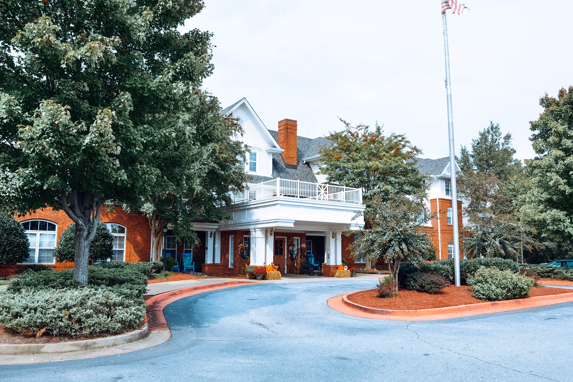 Front entrance of a red-brick senior living building with a covered porte-cochere, circular driveway, trees, and a flagpole.