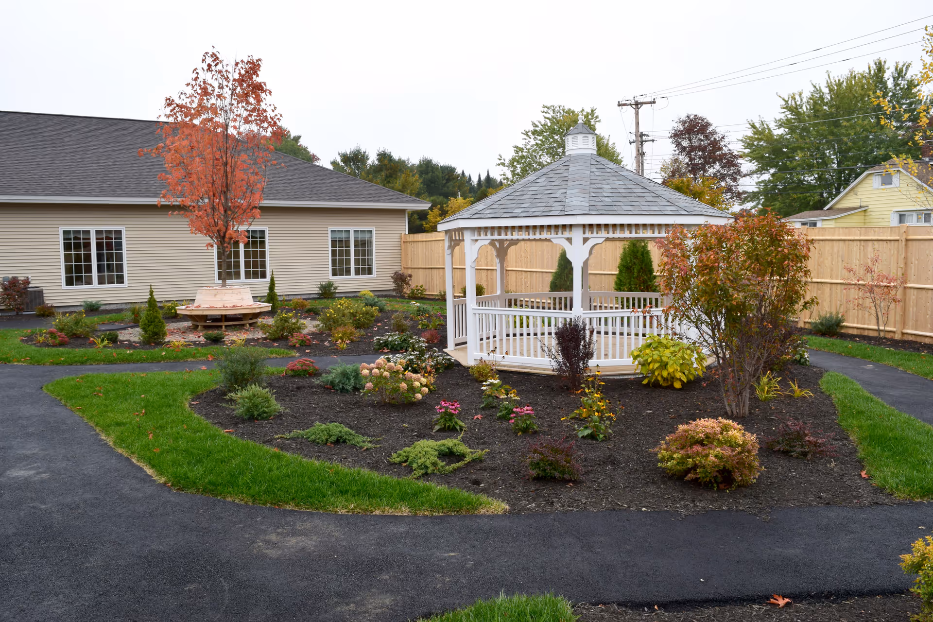 A landscaped courtyard featuring a white gazebo, garden beds, paved walkways, and a beige building in the background.