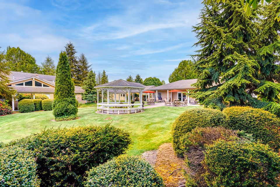 A well-maintained outdoor garden area at The Cottages of Snohomish featuring a white gazebo in the center, surrounded by green grass, bushes, and tall trees. In the background, there are single-story buildings with patios furnished with chairs and red umbrellas under a partly cloudy blue sky.
