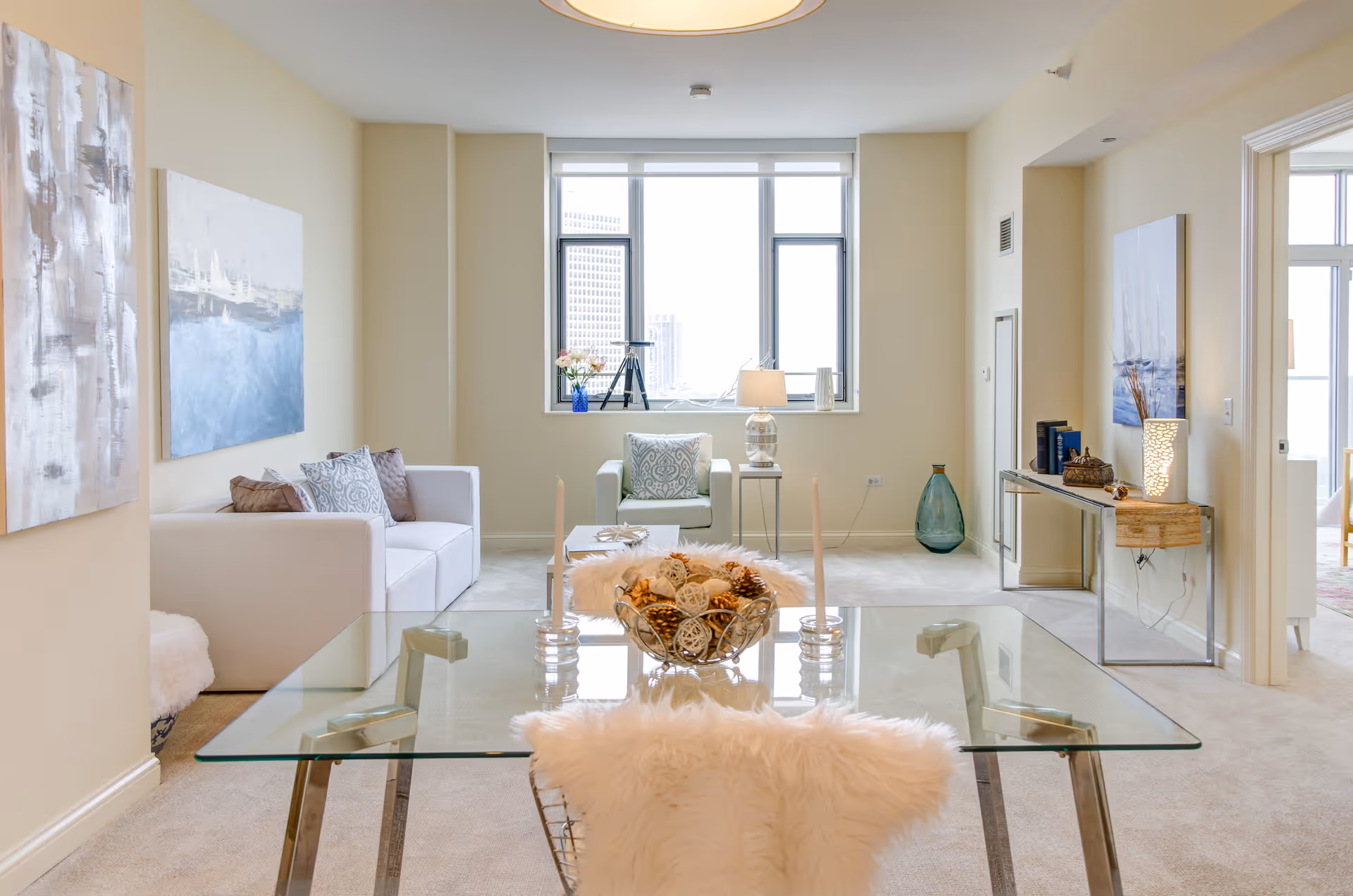 Bright and modern living room with a glass dining table in the foreground, decorated with candles and a centerpiece. The room features a white sofa with patterned pillows, a green armchair near a large window with city views, and light-colored walls adorned with abstract paintings. A console table with decorative items and a lamp is positioned against the right wall.