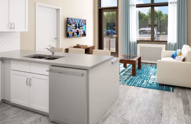 Open-plan interior showing a white kitchen island with sink in the foreground and a living area with sofa, coffee table, wall-mounted TV, and large window/door in the background.