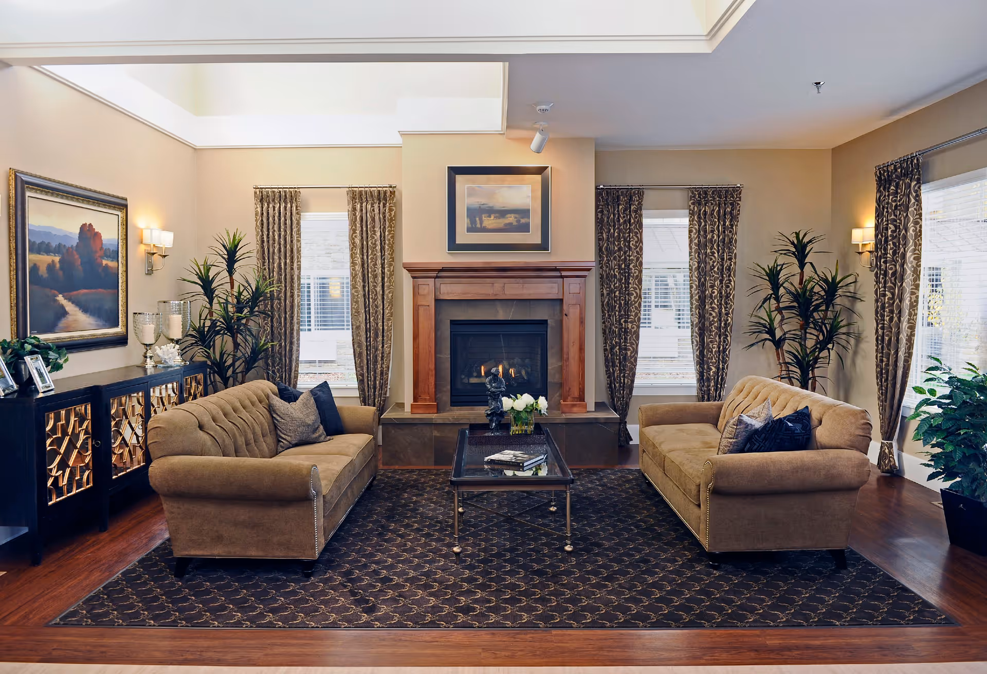 A cozy living room with two beige sofas facing each other across a dark patterned rug. A glass coffee table with books and a flower arrangement sits between the sofas. Behind the table is a fireplace with a wooden mantel and a framed picture above it. The room has three windows with patterned curtains and several potted plants. A sideboard with decorative items and a landscape painting is on the left wall.
