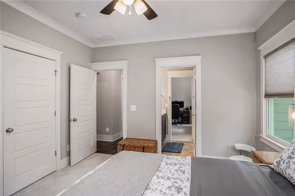 A neutral-toned bedroom with a bed in the foreground, a ceiling fan, two white doors, and an open doorway leading to a bathroom/office area.