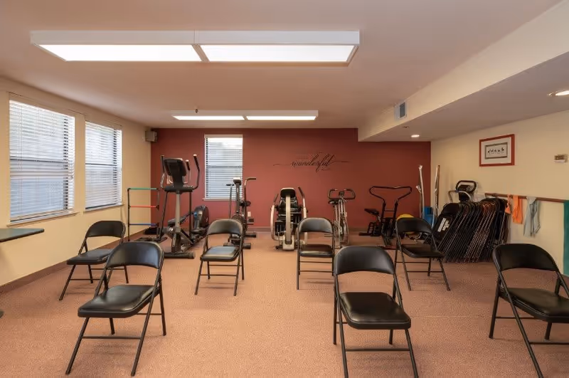Interior fitness/activity room with folding chairs set in rows and several exercise machines against a maroon accent wall.