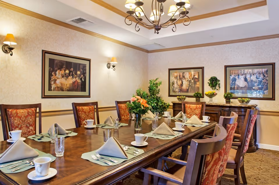 A formal dining room with a long wooden table set for eight people. Each place setting includes a folded beige napkin, a cup and saucer, a glass, and silverware on a green placemat. The chairs have wooden frames with red and gold patterned upholstery. The room is decorated with framed paintings on the walls, wall sconces with yellow lampshades, a chandelier, and a sideboard with plants and decorative items.