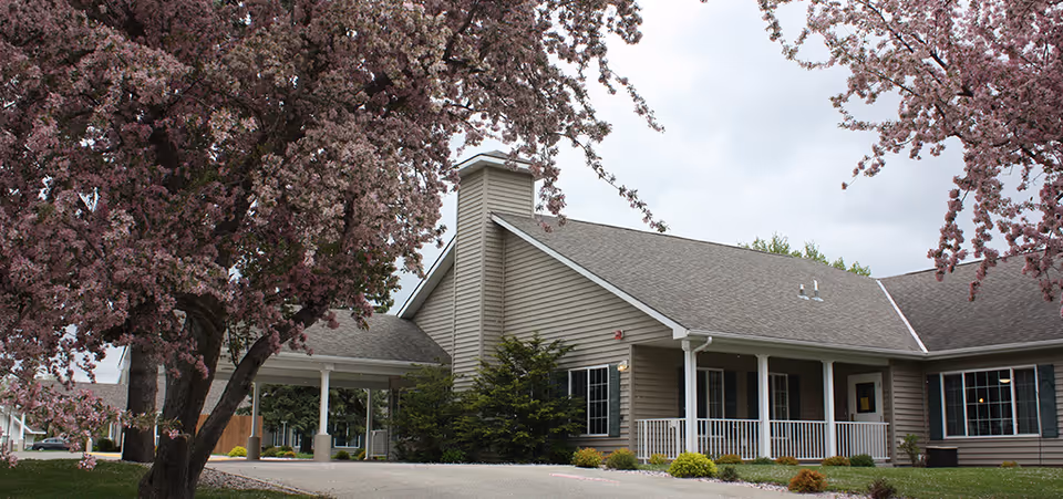 Exterior view of a single-story building with beige siding and a gray roof, surrounded by blooming pink cherry blossom trees and green shrubs under a cloudy sky.