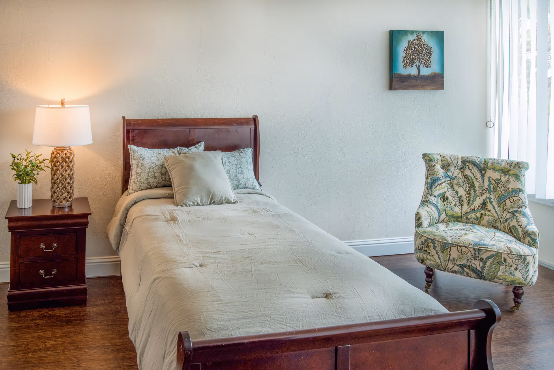 Furnished bedroom with a single wooden bed, nightstand with lamp and plant, and a patterned armchair by a window.