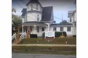 A large, white, multi-story house with a turret and a covered front porch. There is a sign on the lawn that reads 'Wyalusing Valley Retirement And Personal Care Home.' The house is surrounded by a small lawn and some shrubs, with a sidewalk and street in the foreground.