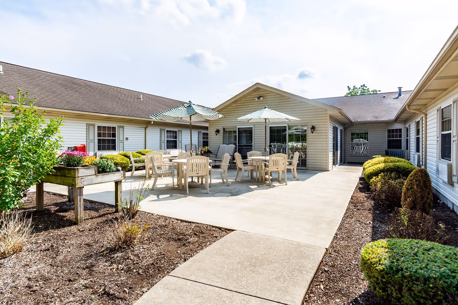 Outdoor patio area at Colonial Courtyard at Tyrone featuring multiple round tables with beige chairs and umbrellas, surrounded by a garden with shrubs and flowers, adjacent to a single-story building with beige siding and multiple windows.