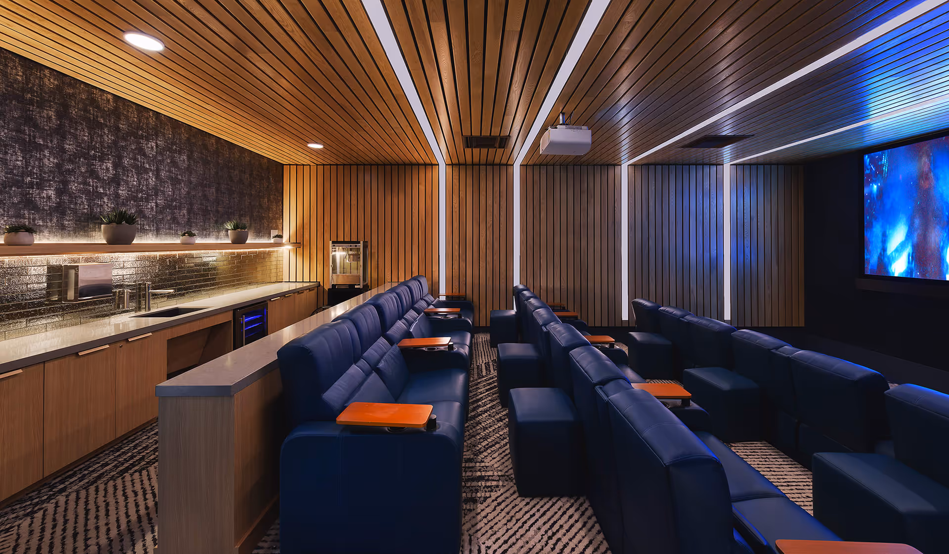 A modern home theater room with rows of blue leather reclining seats equipped with wooden armrest trays. The walls and ceiling are covered with vertical wooden panels, and there is a large screen displaying a colorful image on the right. On the left side, there is a countertop with cabinets below, small potted plants, a popcorn machine, and ambient lighting under a shelf.