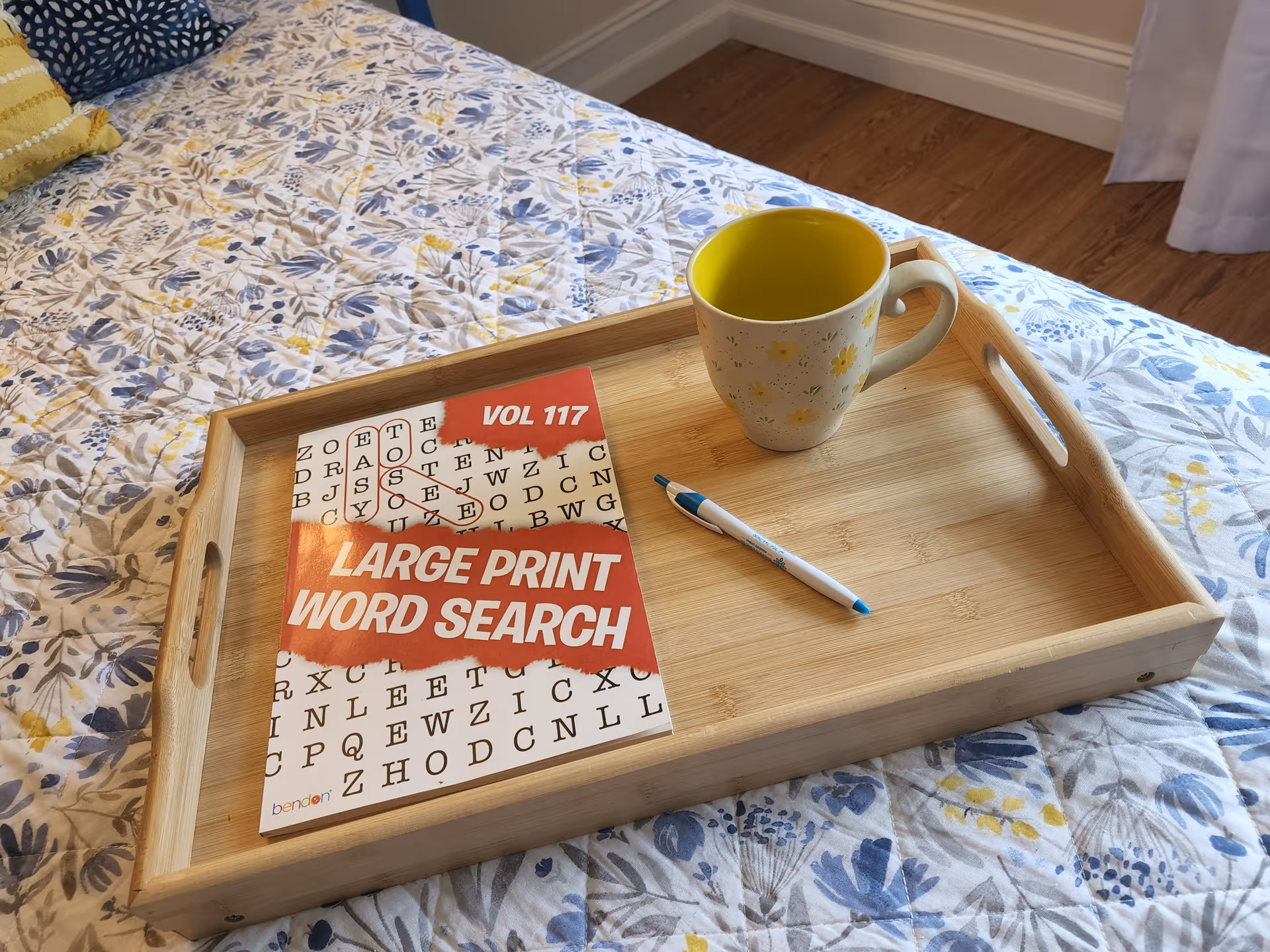 A wooden tray on a floral bedspread holds a 'Large Print Word Search' book, a pen, and a yellow-lined mug.