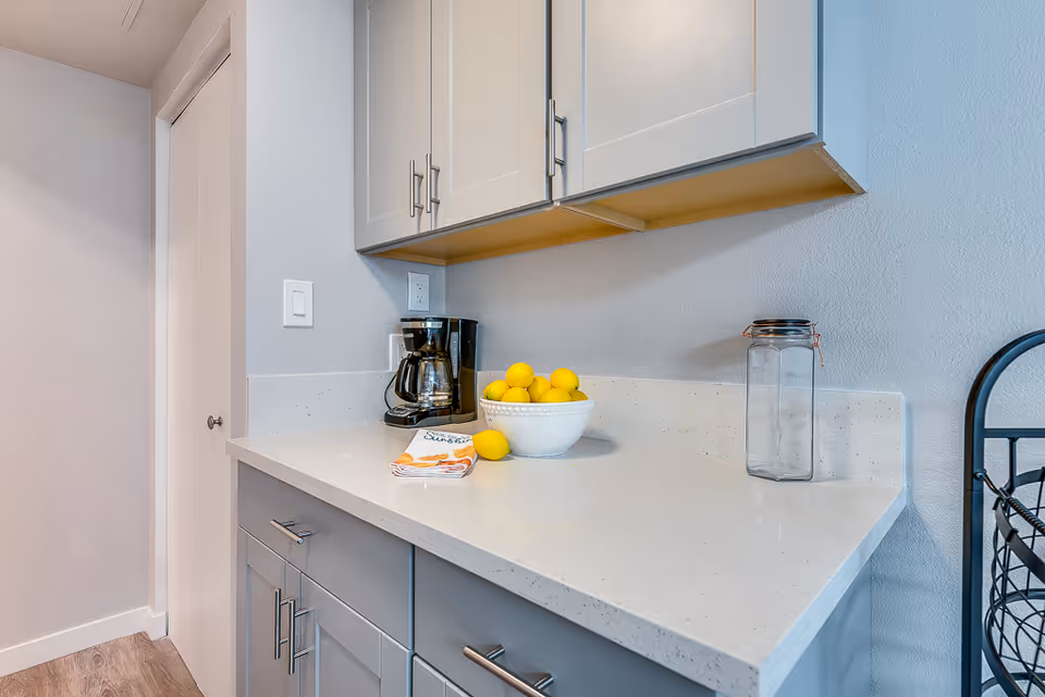A clean kitchen countertop with a bowl of lemons, a coffee maker, a glass jar, and cabinets overhead.
