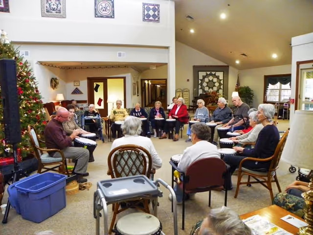 A group of elderly people sitting in a circle in a common room, each holding a drum, participating in a group drumming activity. The room is decorated with a Christmas tree, framed quilt patterns on the walls, and has a cozy, well-lit atmosphere.