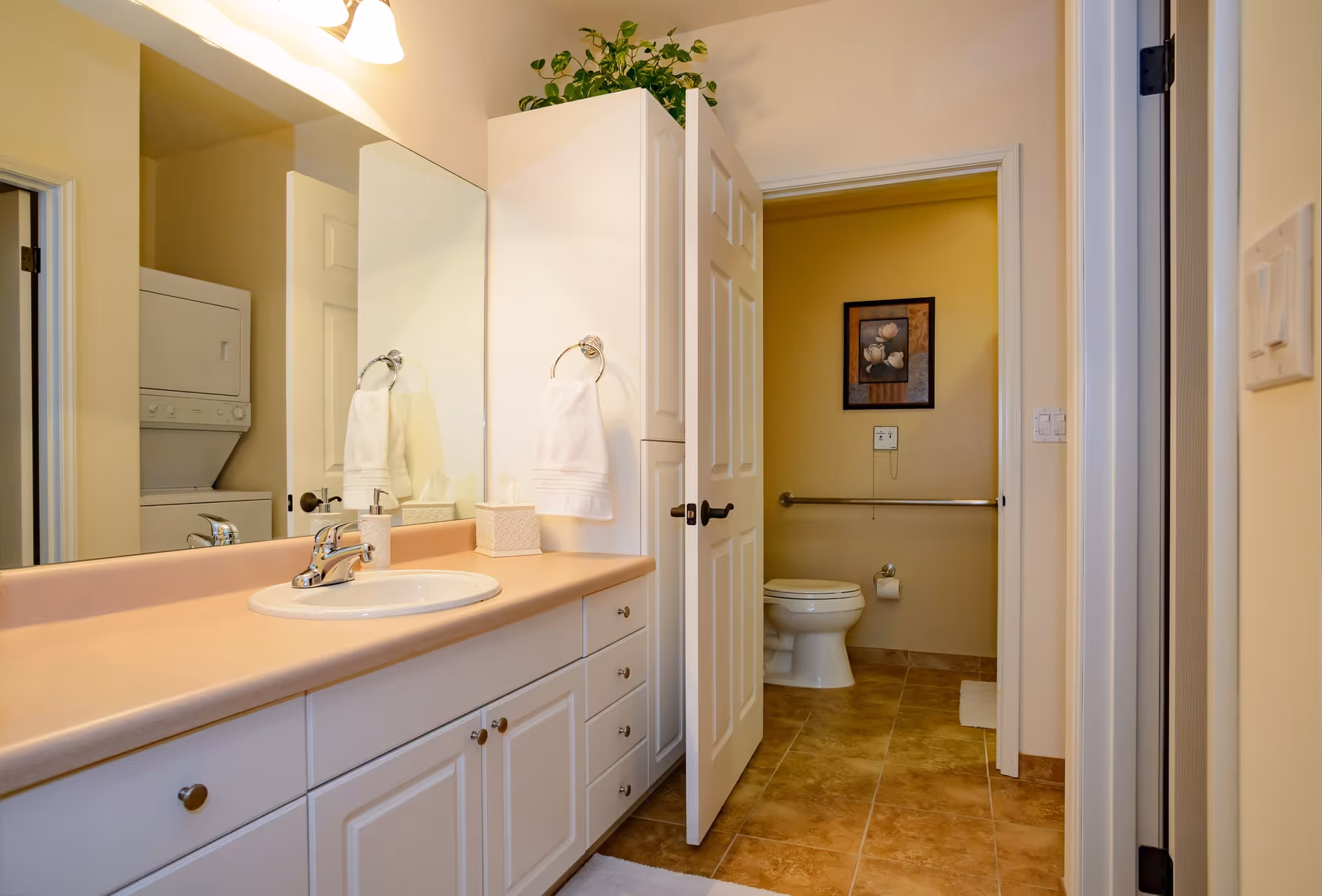 A clean bathroom with a large mirror above a countertop with a sink, soap dispenser, tissue box, and towel ring holding a white towel. The bathroom has white cabinets and drawers below the countertop. Through an open door, a toilet with a grab bar and a framed floral picture on the wall is visible. A stacked washer and dryer unit is seen in an adjacent room.