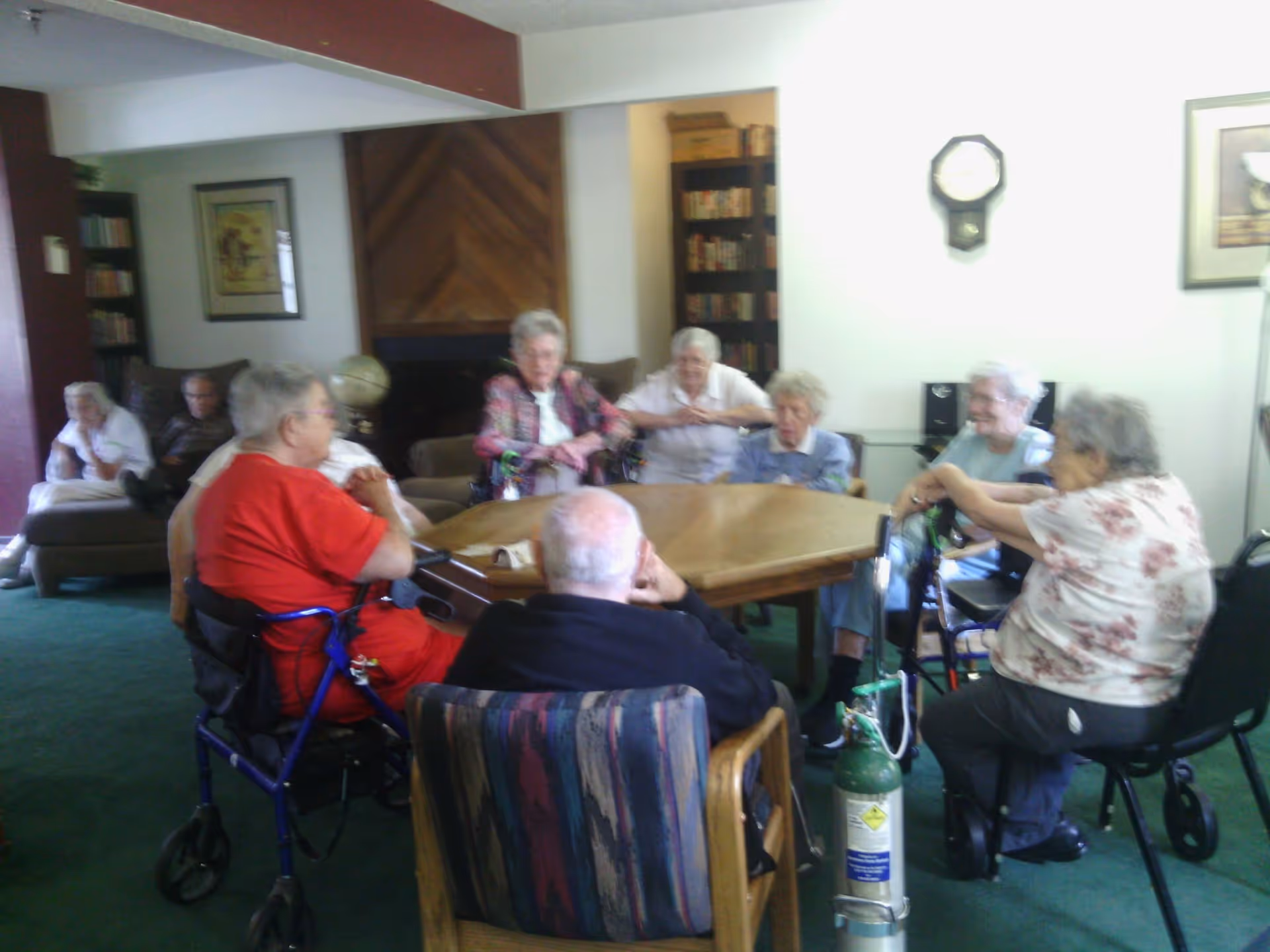A group of elderly people sitting around a wooden table in a room with green carpet. Some are seated in wheelchairs and others in chairs. The room has bookshelves, framed pictures on the walls, and a clock. The atmosphere appears to be a common area or lounge in a senior living facility.
