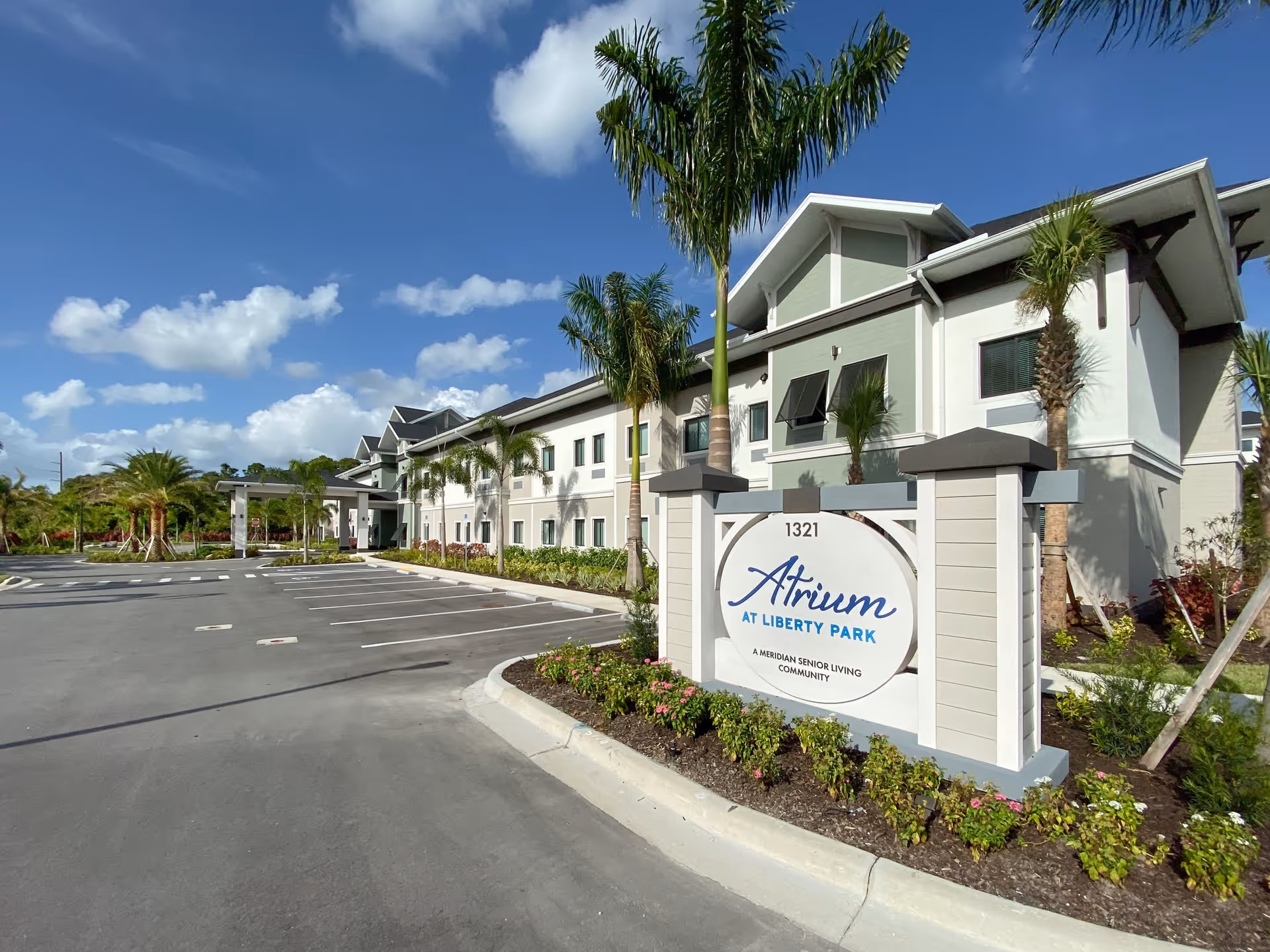 Exterior view of Atrium at Liberty Park senior living community building with a clear blue sky, palm trees, and an empty parking lot in front. A large sign with the facility name is visible near the entrance.