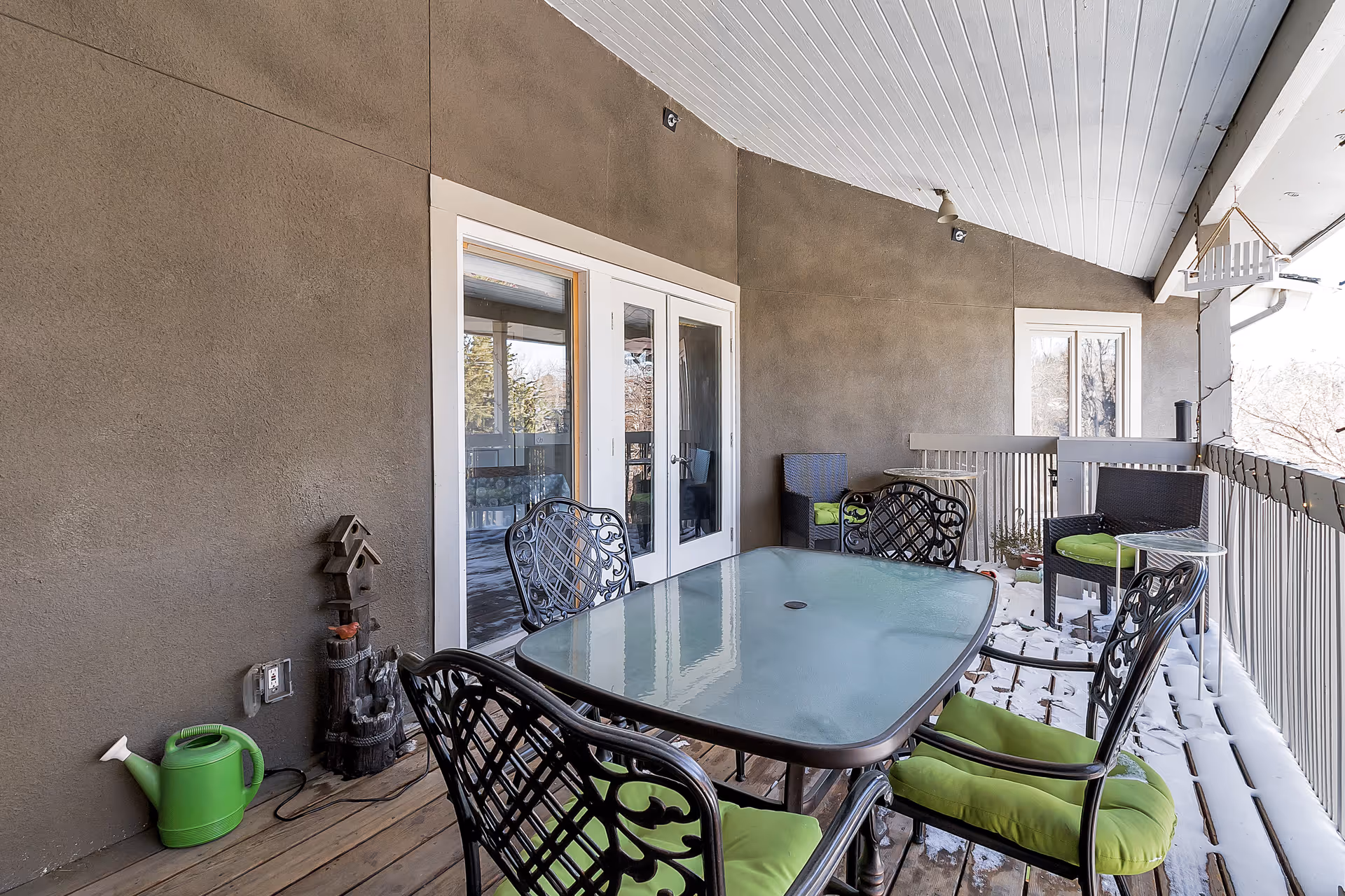 Covered outdoor patio area with a glass-top table surrounded by four black metal chairs with green cushions. There are additional seating chairs along the wall, a green watering can on the wooden floor, and a small birdhouse decoration. Snow is visible on the floor and railing outside the patio.