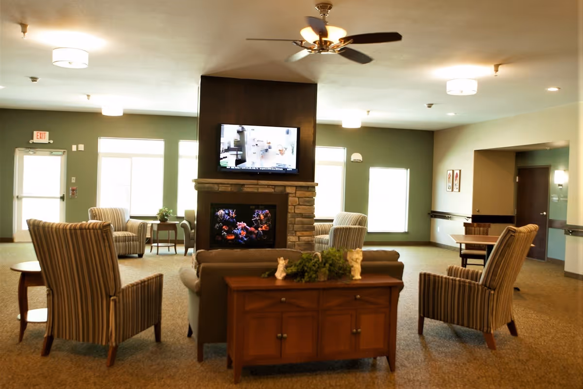 A spacious living room area in a senior living facility featuring a central stone fireplace with a TV mounted above it. The room has multiple striped armchairs and a sofa arranged around the fireplace. There is a wooden console table with decorative items behind the sofa. The room is well-lit with ceiling lights and natural light from windows.