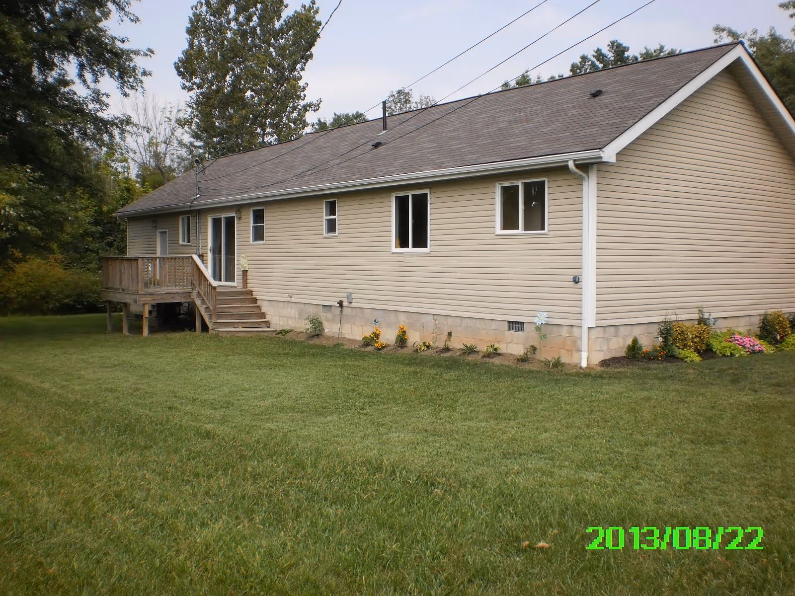 Side view of a single-story beige house with a small wooden deck and stairs leading to a door. The house has several windows and is surrounded by a well-maintained lawn with some small plants and flowers near the foundation. Trees are visible in the background under a partly cloudy sky.