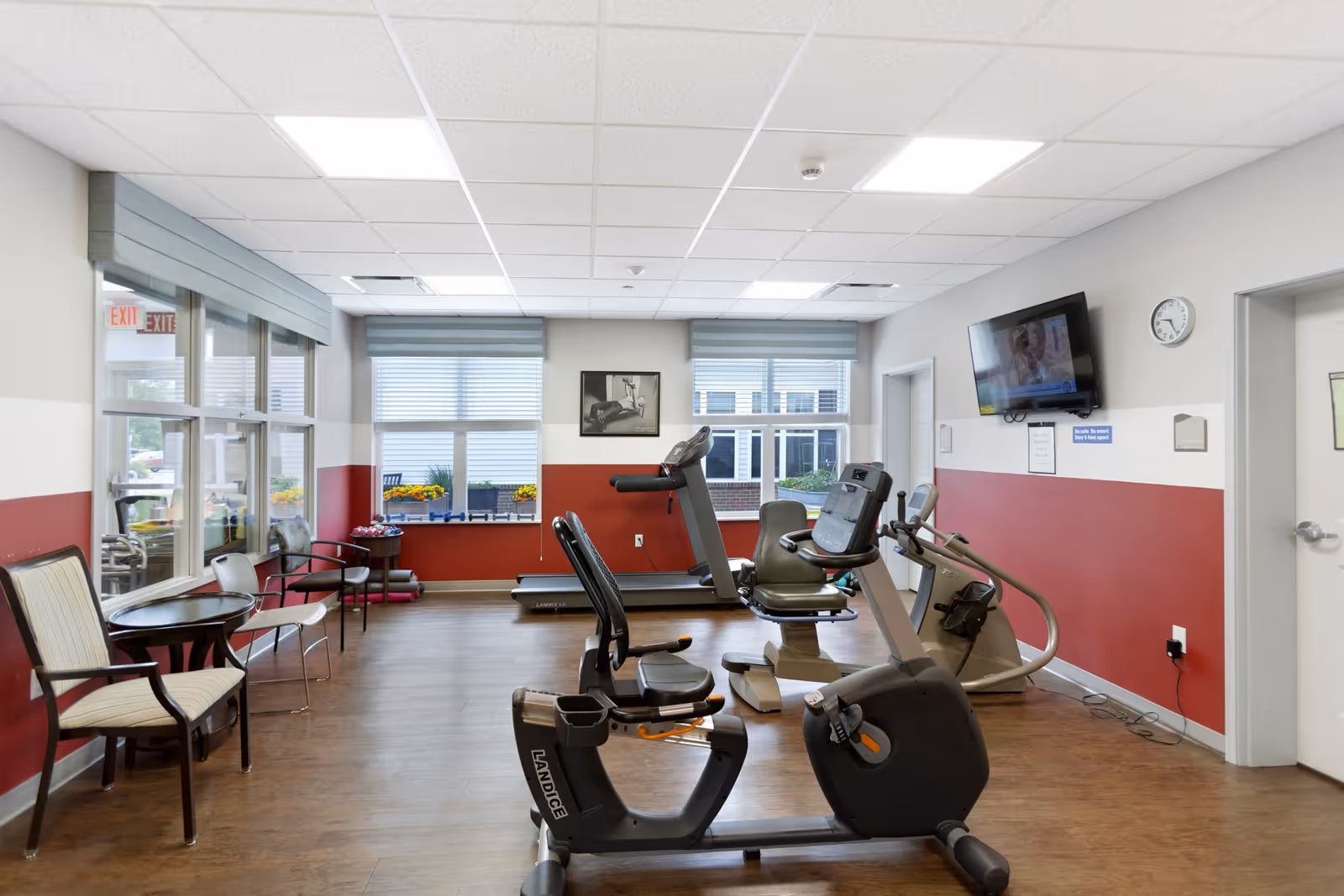 A fitness room in an assisted living facility with exercise equipment including a recumbent bike, treadmill, and elliptical machine. The room has large windows with blinds, a wall-mounted TV, a clock, and a few chairs and tables along the left side. The walls are painted white and red, and the floor is wood.