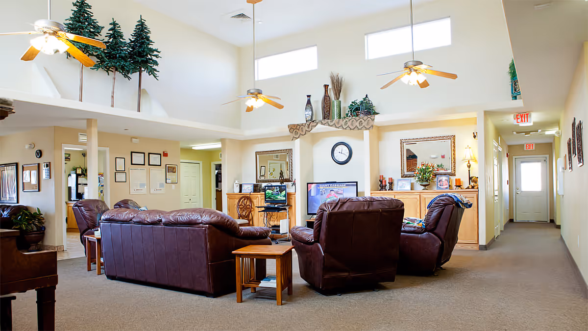 A bright and spacious living room area in a senior living facility with high ceilings, ceiling fans, and large windows near the ceiling. The room is furnished with brown leather sofas and armchairs arranged around a small wooden table. There is a TV on a stand against the wall, a clock above it, and decorative items including plants and framed pictures on a wooden cabinet. The walls are light-colored, and there is a hallway leading to a door with exit signs visible.