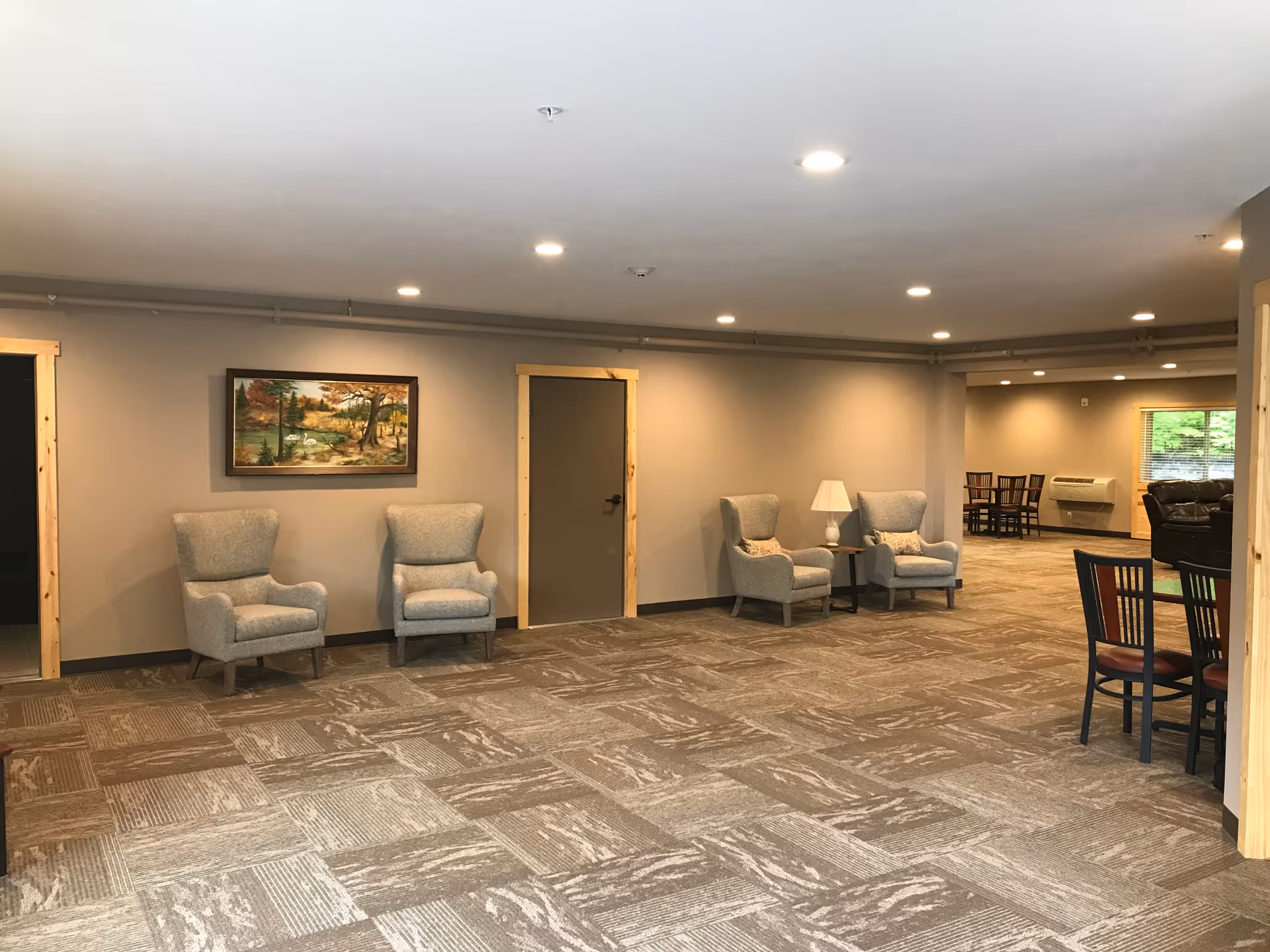 A spacious common area in an assisted living facility with four gray upholstered armchairs arranged along the walls, a small table with a lamp between two of the chairs, a framed landscape painting on the wall, and a dining area with tables and chairs visible in the background. The room has recessed lighting and carpeted flooring with a patterned design.