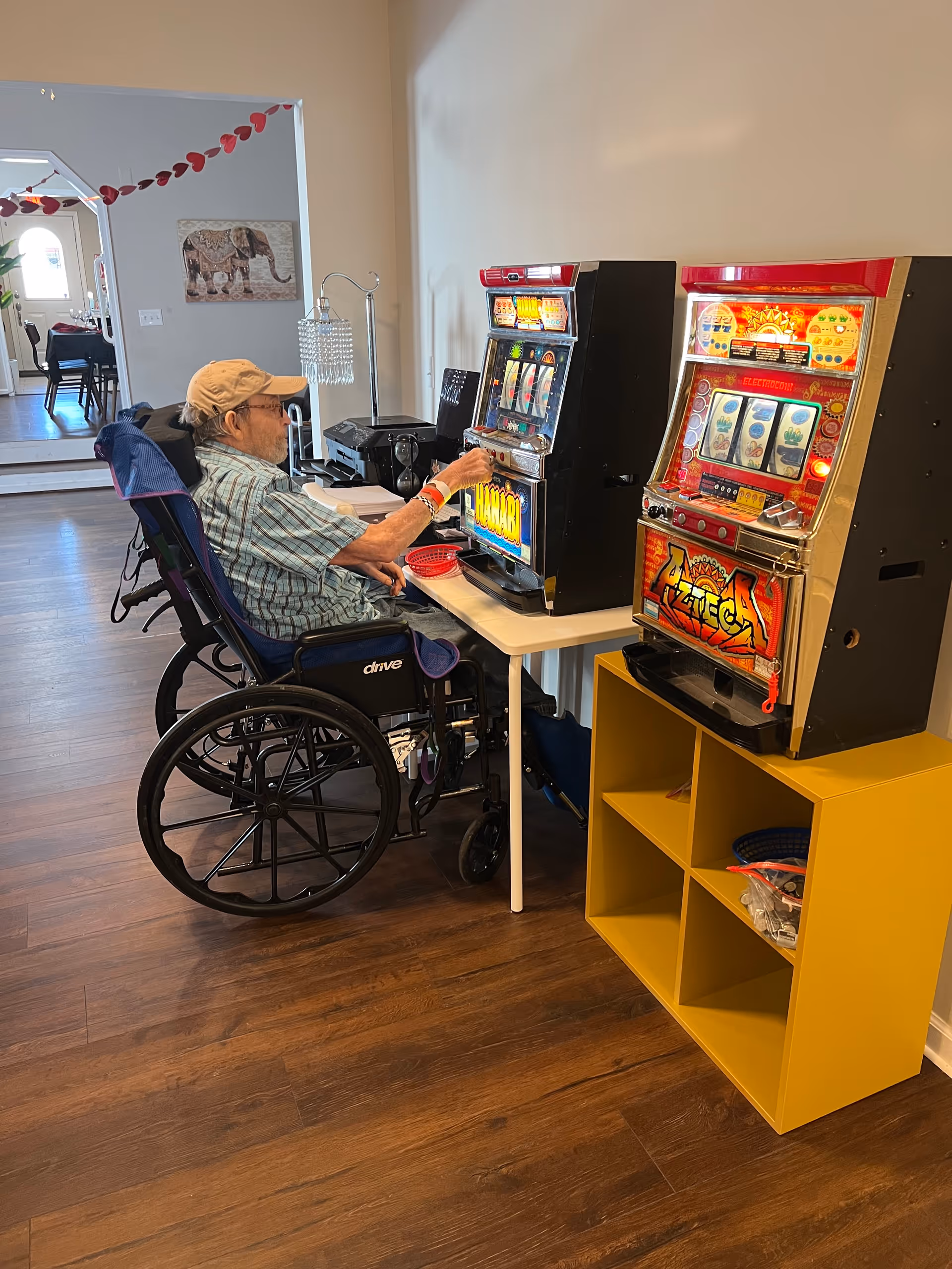 An elderly man in a wheelchair playing on a slot machine in a room with wooden flooring. There are two slot machines placed on a white table and a yellow shelving unit. In the background, there is a hallway with a dining area and a wall decoration of an elephant.
