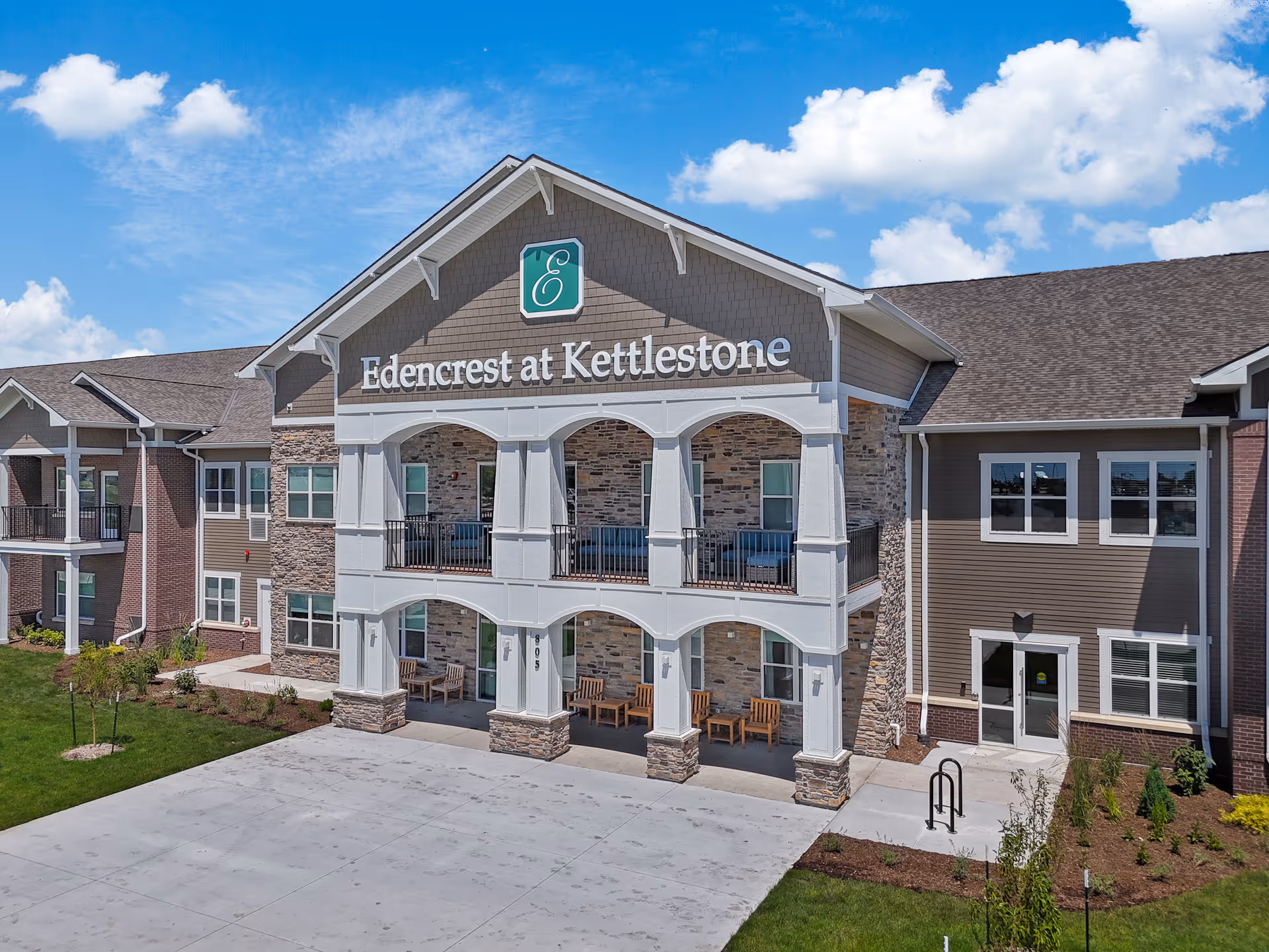 Exterior front view of Edencrest at Kettlestone senior living facility showing a two-story building with a covered porch area, stone and siding facade, outdoor seating, and a clear blue sky with some clouds.