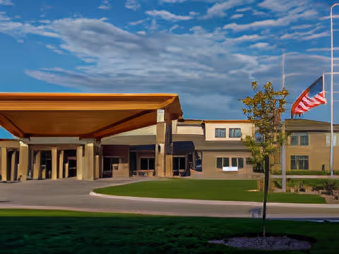 Exterior view of Brookestone Gardens facility with a covered entrance, a flagpole displaying the American flag, a small tree, and a well-maintained lawn under a partly cloudy sky.