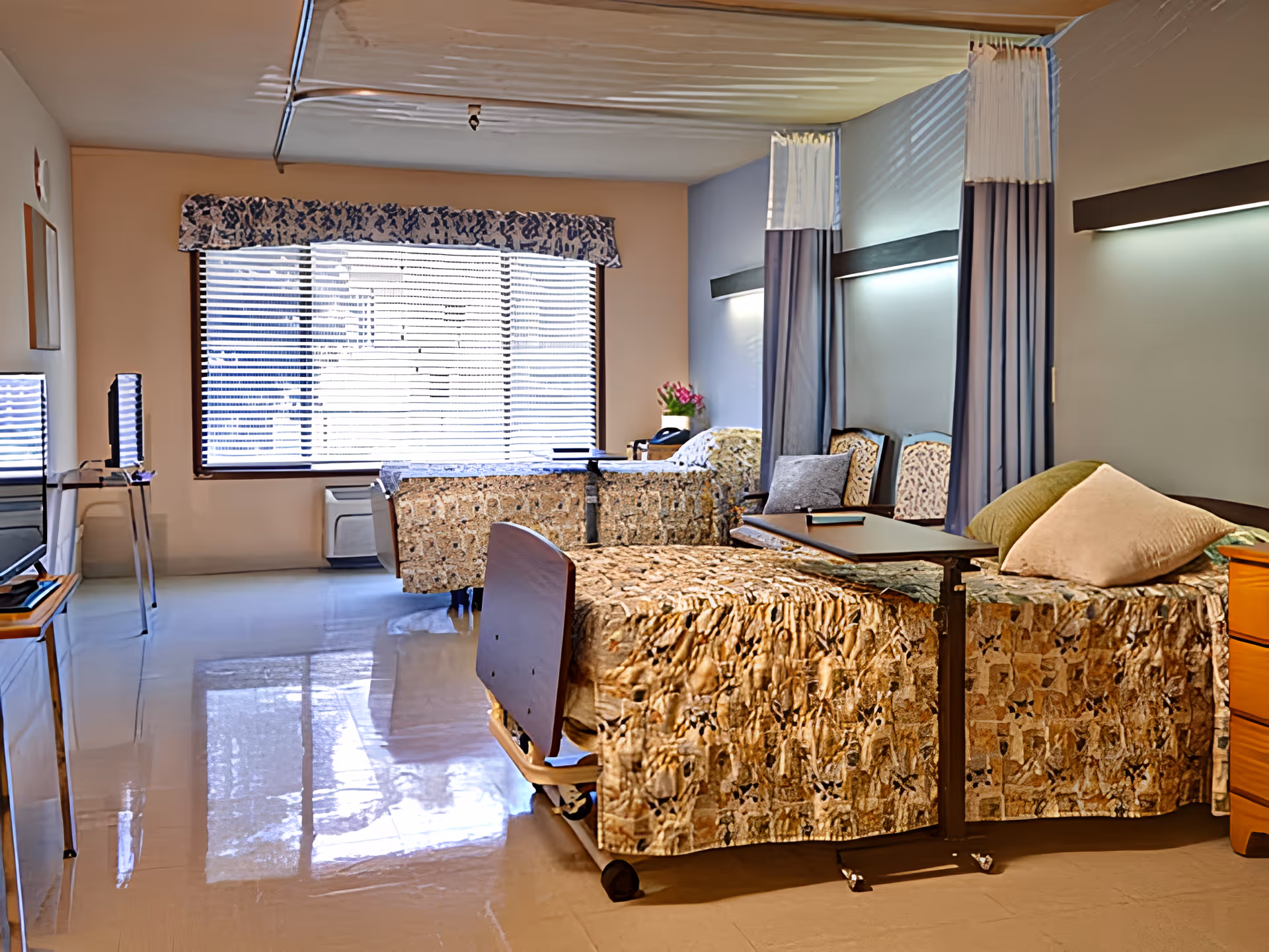 A shared patient room in a transitional care facility with two hospital beds covered in patterned bedspreads. Each bed has a bedside table and an overbed table. There are two chairs against the wall with privacy curtains hanging above them. A large window with blinds and a floral valance lets in natural light. The room has a polished floor and soft lighting.