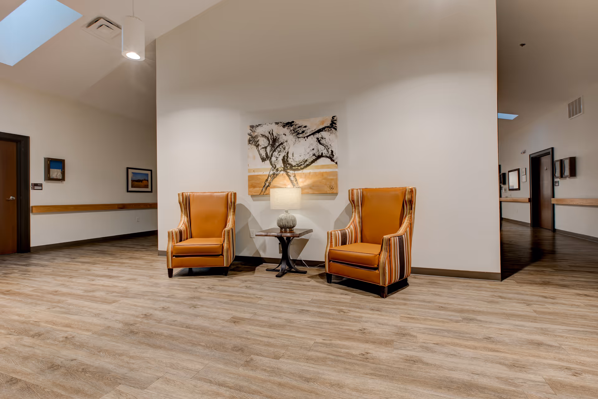 Two orange wingback chairs flank a small table with a lamp beneath a horse painting in a clean interior hallway.