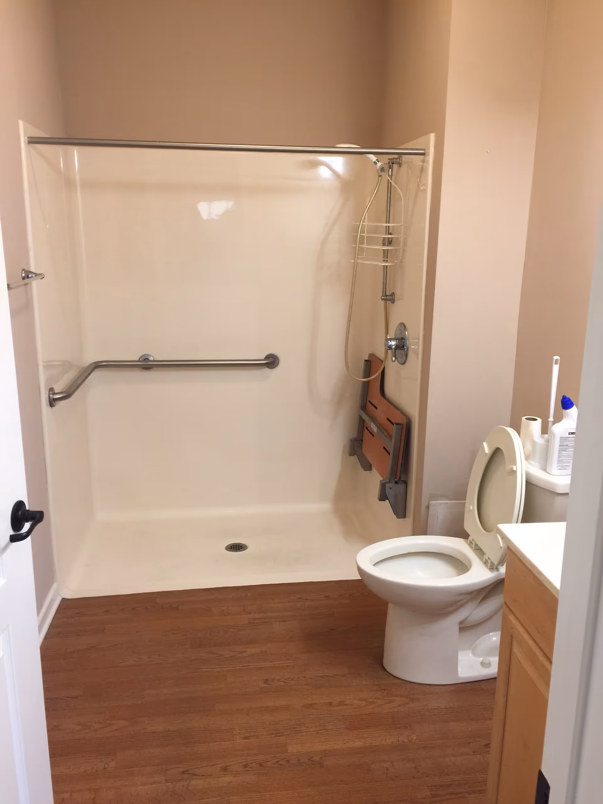 A bathroom with a walk-in shower featuring a grab bar and a fold-down shower seat. There is a toilet with the lid open next to a vanity with a white countertop. The floor has wood-like flooring and the walls are painted beige.