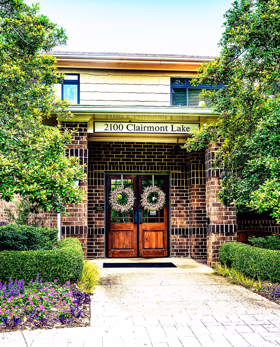 Front entrance of a brick building with double wooden doors decorated with two floral wreaths. Above the entrance is a sign that reads '2100 Clairmont Lake'. The entrance is framed by green bushes and trees on both sides, with a paved walkway leading up to the doors.