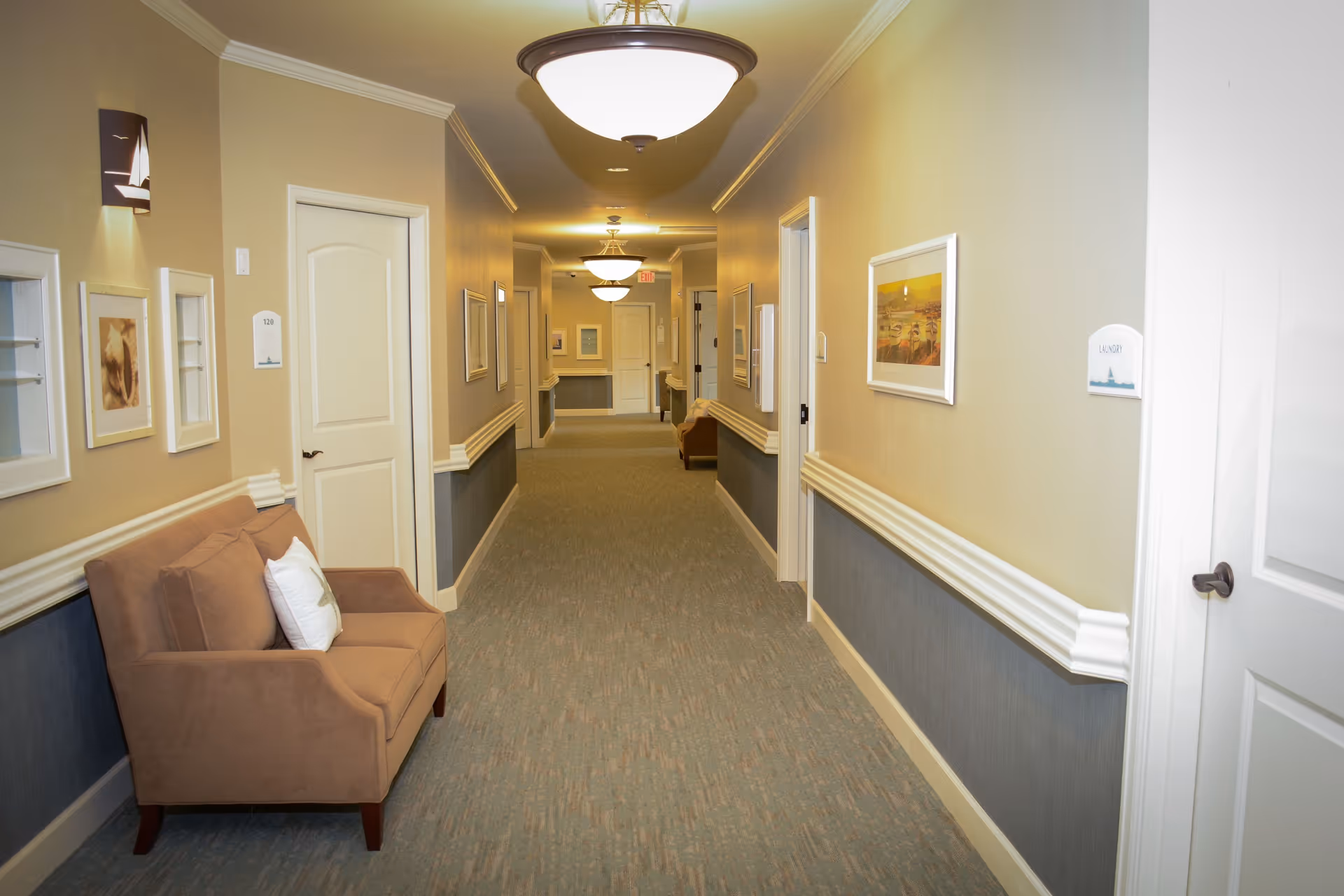 A well-lit hallway in a senior living facility with beige and blue walls, carpeted floor, several closed white doors, framed artwork on the walls, and a brown cushioned bench with pillows on the left side.