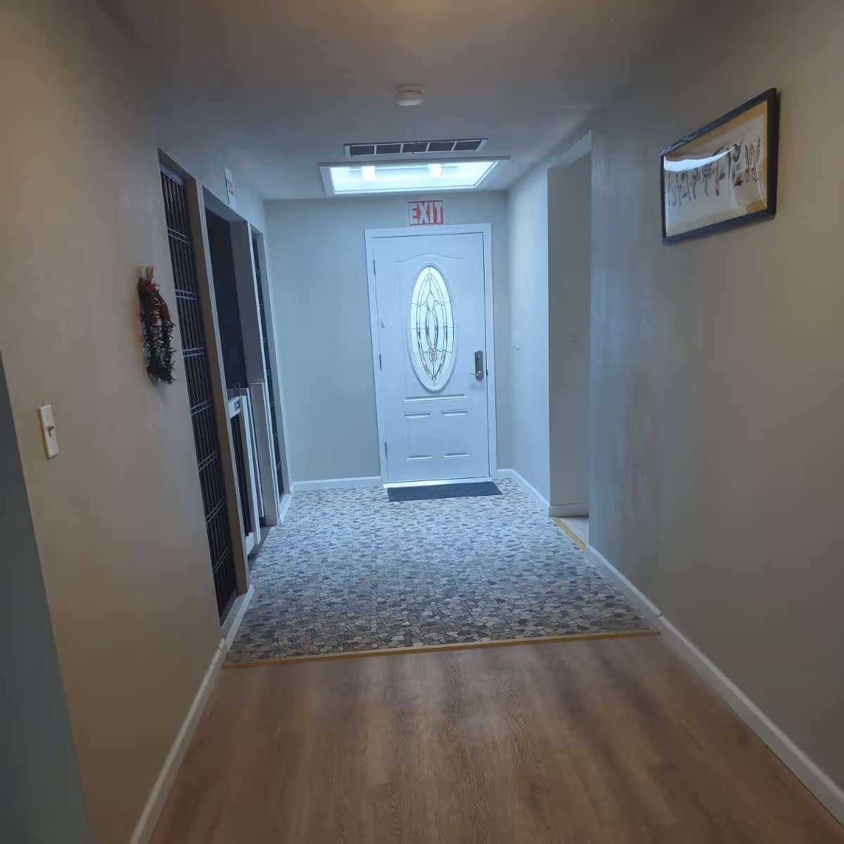 A hallway with light-colored walls and wood flooring leading to a white door with an oval glass window. Above the door is a red EXIT sign and a skylight. There is a patterned rug in front of the door, a small decorative wreath on the left wall, and a framed picture on the right wall.