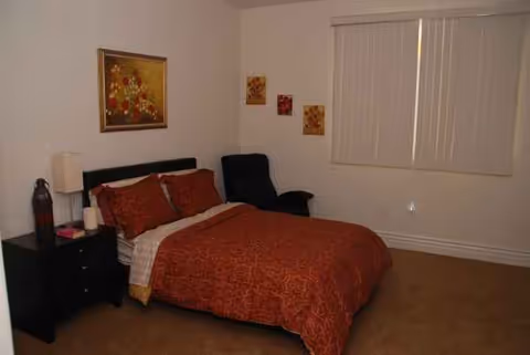 A furnished bedroom with a red quilted bed, nightstand, chair, and window blinds.