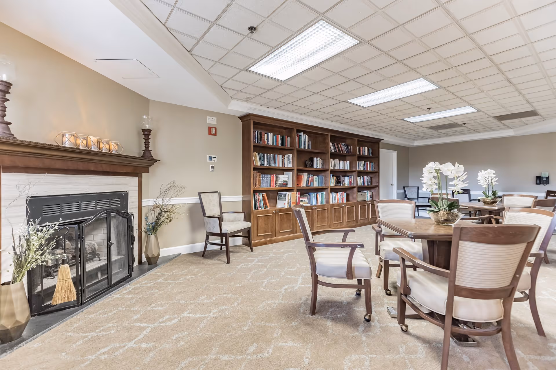A cozy common area with a fireplace on the left, decorated with candles and vases with plants. There is a large wooden bookshelf filled with books against the back wall. Several wooden chairs with cushioned seats surround round tables, each table adorned with a vase of white flowers. The room has a beige carpet with a subtle pattern and a white ceiling with fluorescent lighting.