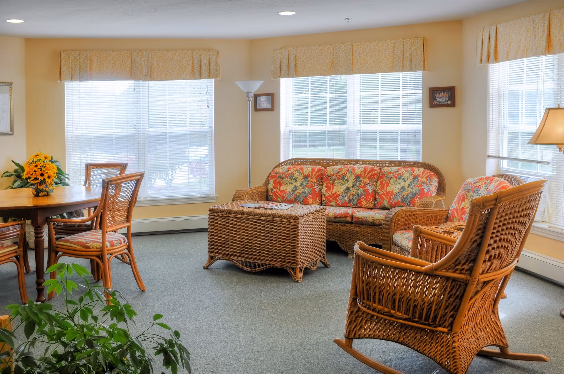 A bright and cozy living room area with wicker furniture including a floral-patterned sofa, two armchairs, and a coffee table. There is a wooden dining table with chairs on the left side, a floor lamp near the windows, and several large windows with blinds and valances letting in natural light. A potted plant is visible in the foreground.