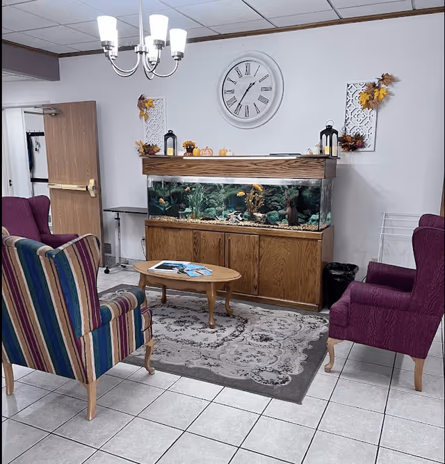 A cozy sitting area with two upholstered armchairs, one striped and one solid purple, facing a wooden coffee table on a patterned rug. Behind the table is a large fish tank with various decorations inside, set against a white wall with a large round clock and autumn-themed wall decor. A chandelier with five lights hangs from the ceiling.