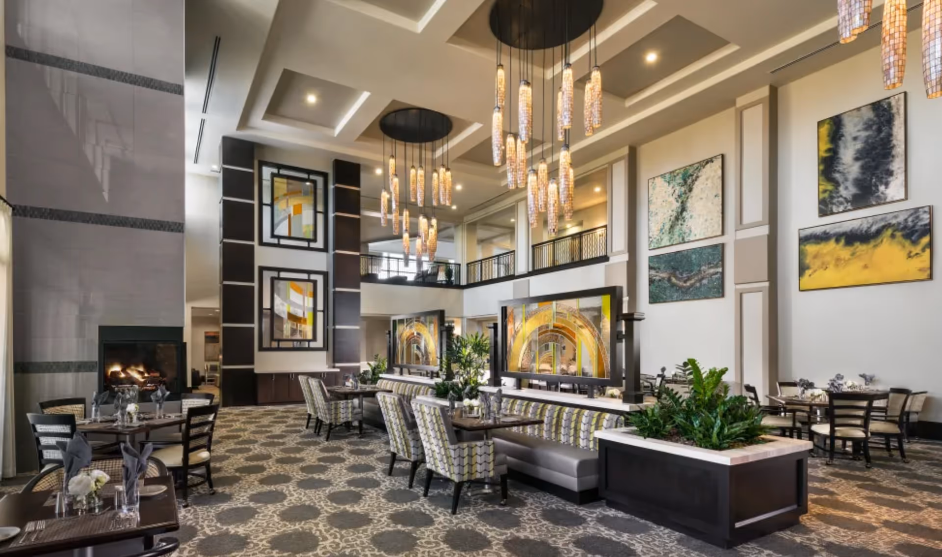 Spacious upscale communal dining and lounge area with high ceilings, hanging pendant lights, multiple tables and seating, a fireplace, and large wall art.