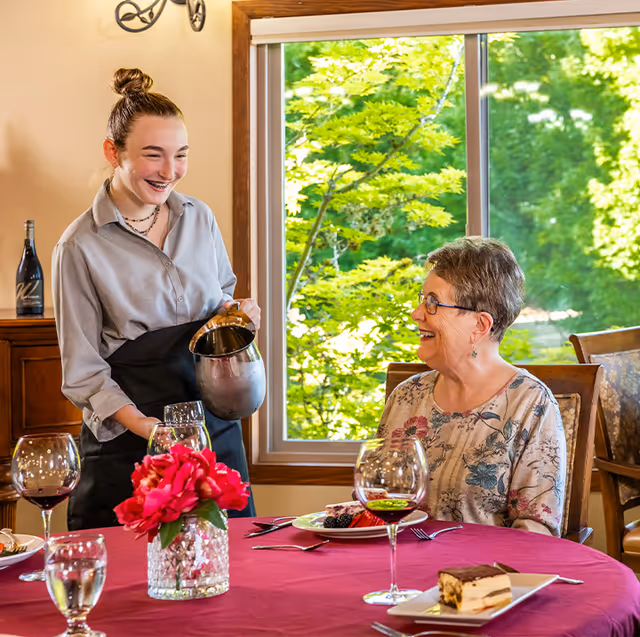 A young female server with a bun hairstyle and braces is smiling and pouring a drink from a metal pitcher for an elderly woman with short gray hair and glasses, who is seated at a dining table with a red tablecloth. The table has wine glasses, a water glass, a vase with red flowers, and plates with dessert. A large window behind them shows green trees outside.