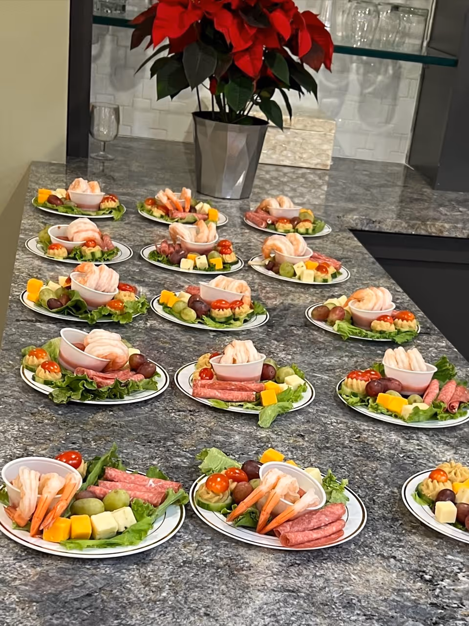 Multiple plates arranged on a granite countertop, each plate containing shrimp cocktail, slices of salami, cubes of cheese, grapes, cherry tomatoes, and lettuce. In the background, there is a silver pot with a red poinsettia plant and glass shelves with glassware.
