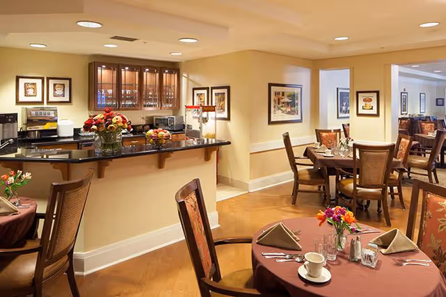 A warm and inviting dining room with round tables covered in burgundy tablecloths, each set with napkins, cups, glasses, and silverware. Chairs with patterned upholstery surround the tables. In the background, there is a counter with a black countertop, decorated with flower arrangements and kitchen appliances. The walls are adorned with framed artwork, and the room has soft, recessed lighting.