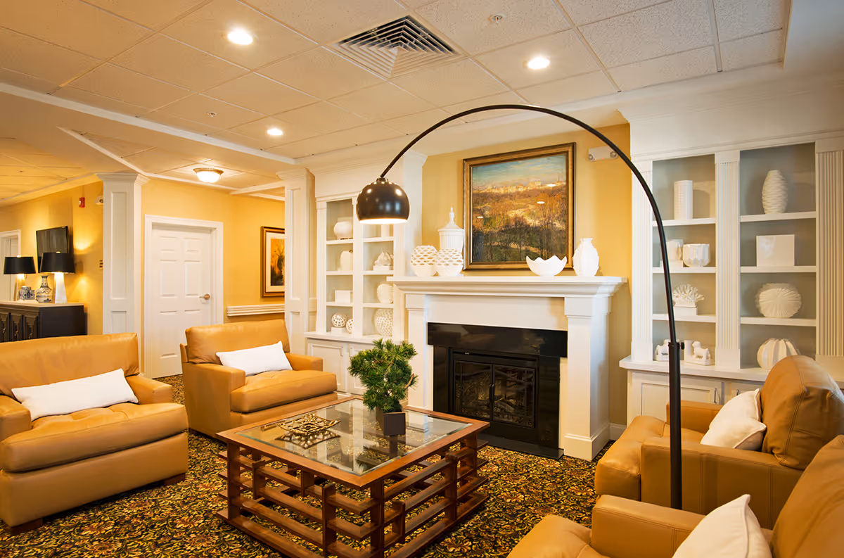 A cozy living room area in an assisted living facility featuring tan leather armchairs with white pillows arranged around a glass-top wooden coffee table. The room has a patterned carpet, a white fireplace with decorative white vases on the mantel, built-in shelves with white decorative items, a large curved floor lamp, and a framed landscape painting above the fireplace. The walls are painted a warm yellow color.