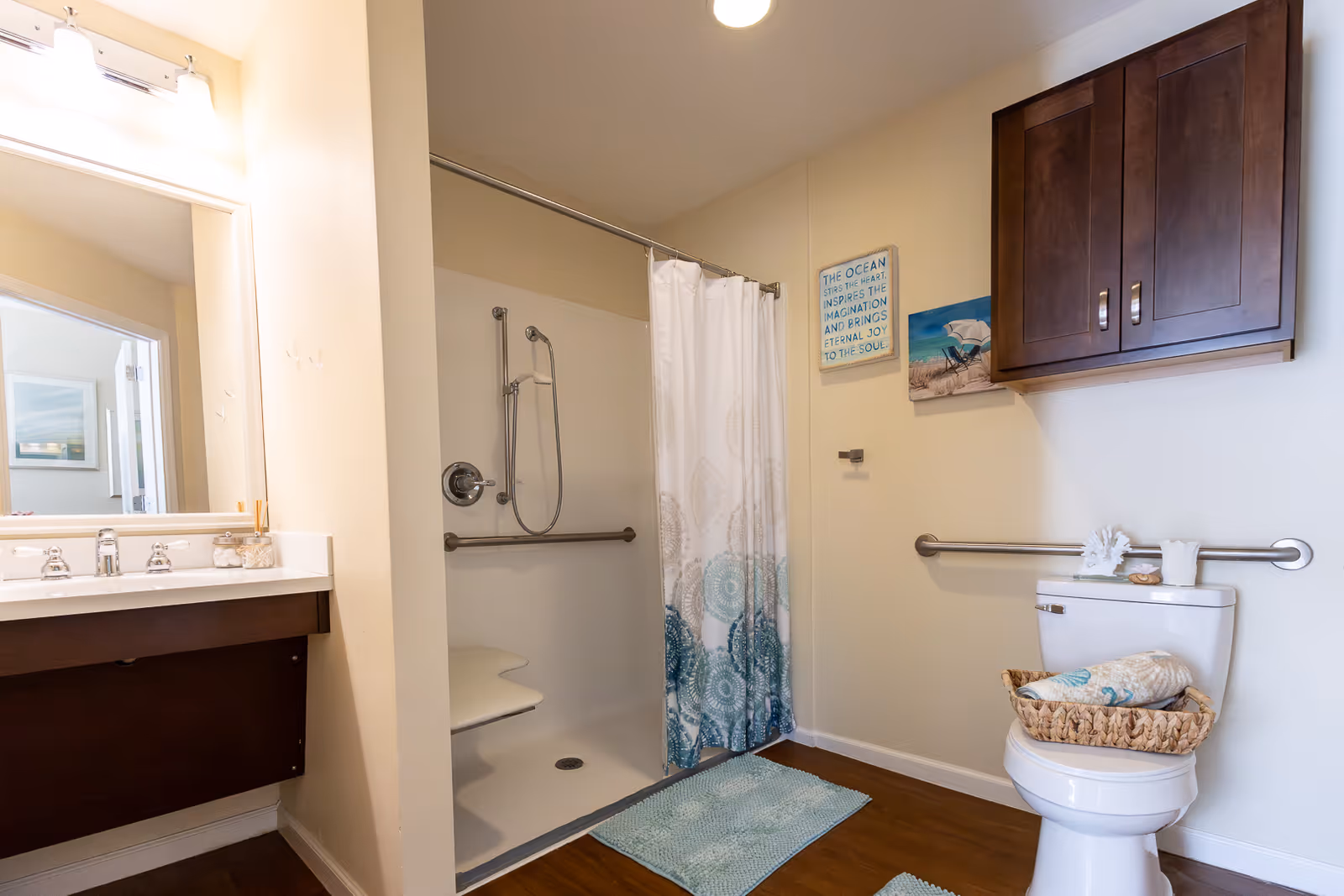 A bathroom with a walk-in shower featuring a white and teal patterned shower curtain, a white toilet with a basket on top containing towels, a wooden cabinet mounted on the wall above the toilet, and a sink with a large mirror and lights above it. The walls are light-colored and there are two blue-green bath mats on the wooden floor.